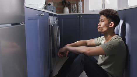 hombre biracial pensativo sentado en el suelo de la cocina, apoyado en el mostrador