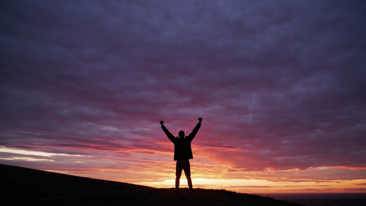 Silhouette of a person with raised arms against a vibrant sunset sky, captured from a low-angle