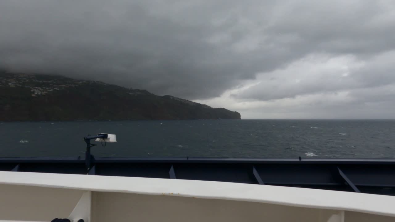 Blick vom Fährdeck beim Segeln nahe der zerklüfteten Klippen der Insel Madeira unter dramatisch bewölktem Himmel. Dunkle Sturmwolken über dem Atlantischen Ozean nahe Funchal, Portugal.