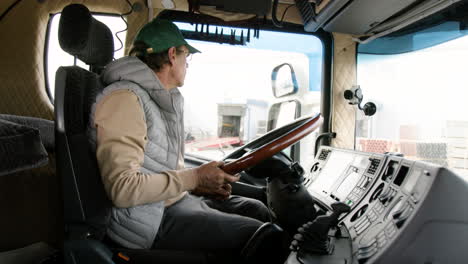 side view of older worker wearing cap and vest driving a truck in a logistics park 2