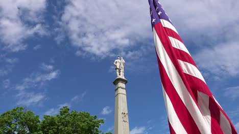 civil war monument statue on memorial day in a cemetery under a cloudy blue sunny sky and the american flag waving on a windy day