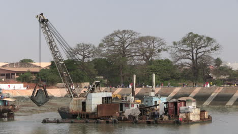 Ein-Großer-Bagger,-Der-Den-Schlamm-Im-Hafen-Von-Bandim-Reinigt-Oder-Vertieft,-Mit-Drei-Großen-Affenbrotbäumen-Im-Hintergrund