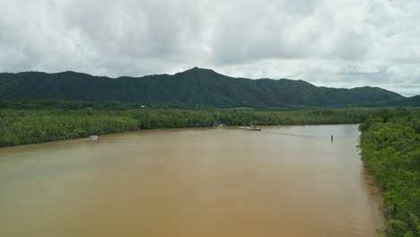 Brown-river-channel-landscape-aerial-drone-fly-forested-Australian-rainforest,-ferry-arrives-to-a-hidden-port,-green-mountain-skyline-background