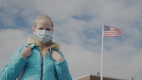 teen in a protective mask stands against the background of a school and a us flag.