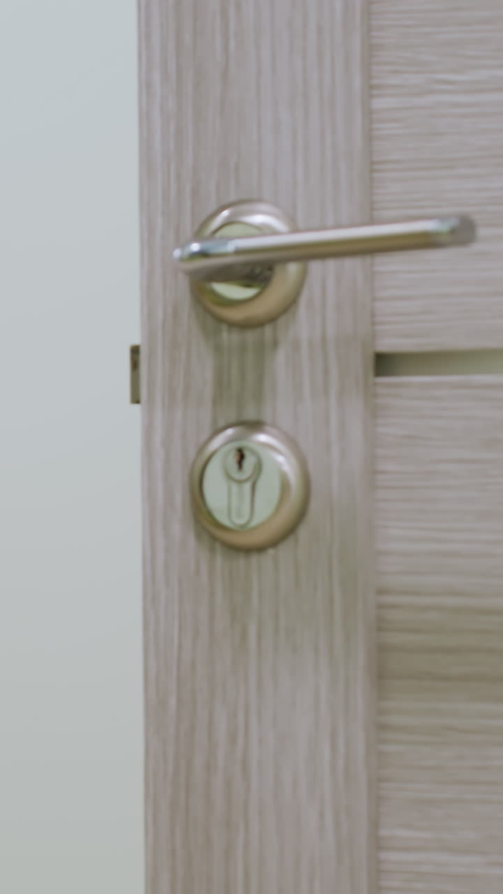 Primer plano de una mujer blanca abriendo una puerta de madera en el pasillo de un apartamento, con detalle del pomo y el ojo de la cerradura. Lleva un jersey gris, es propietaria y examina la cerradura antes de entrar con cautela en una vivienda de ambiente tranquilo.