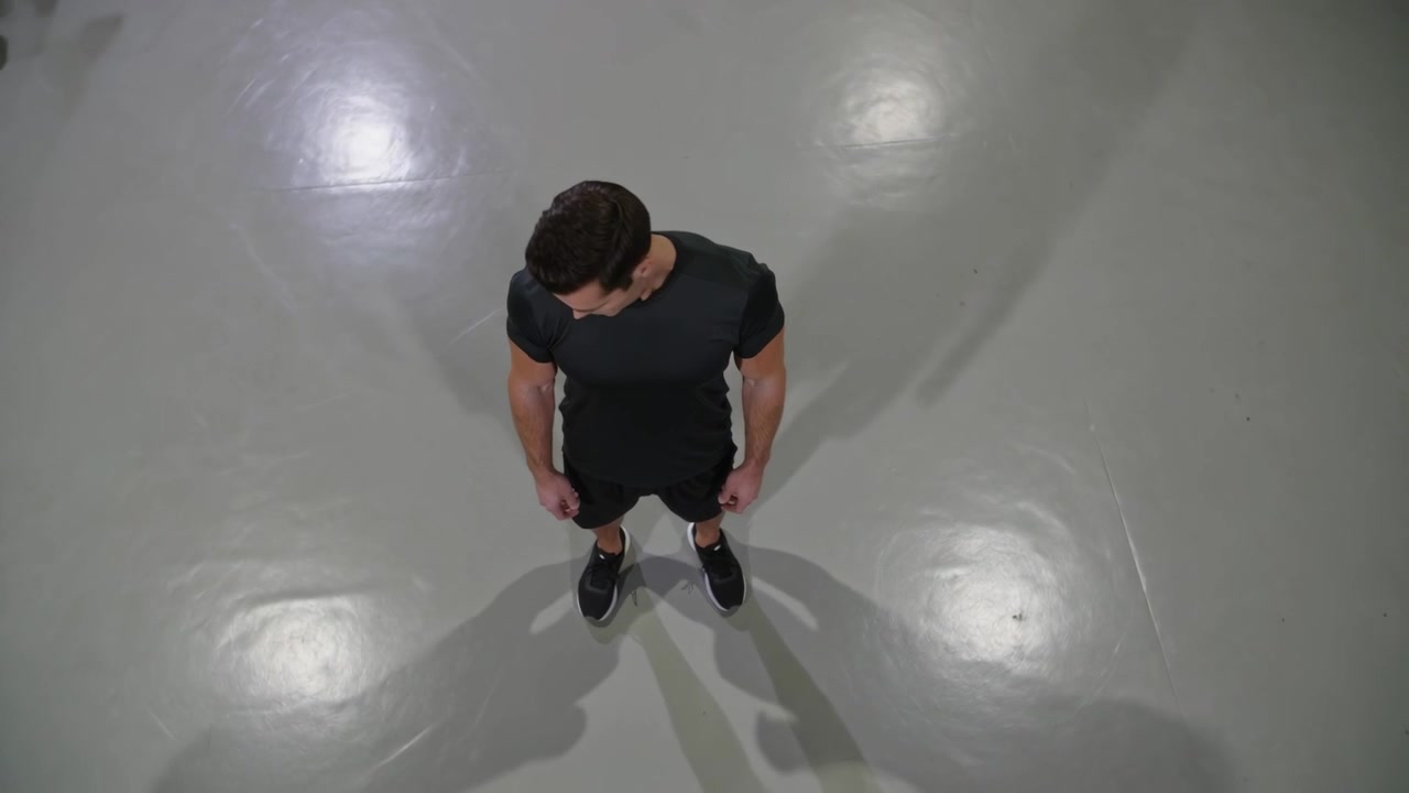 Uomo atletico vestito di scuro si gira, scrutando il pavimento grigio mentre la telecamera registra, mostrando scarpe e ombre.