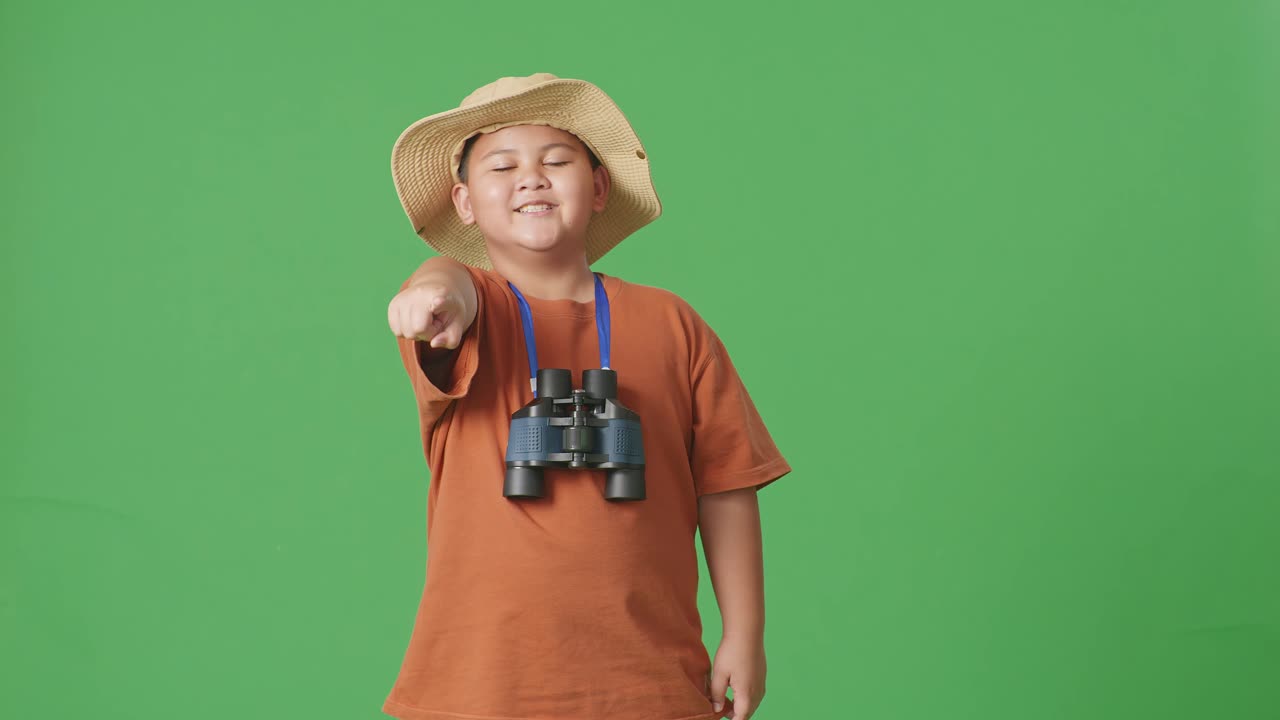 모자와 안경을 쓴 아시아 관광 소년이 웃으며 가을 만지고 녹색 스크린 배경에 서서 당신을 가리키고 있습니다. 소년 연구자는 여행 관광을 조사합니다.