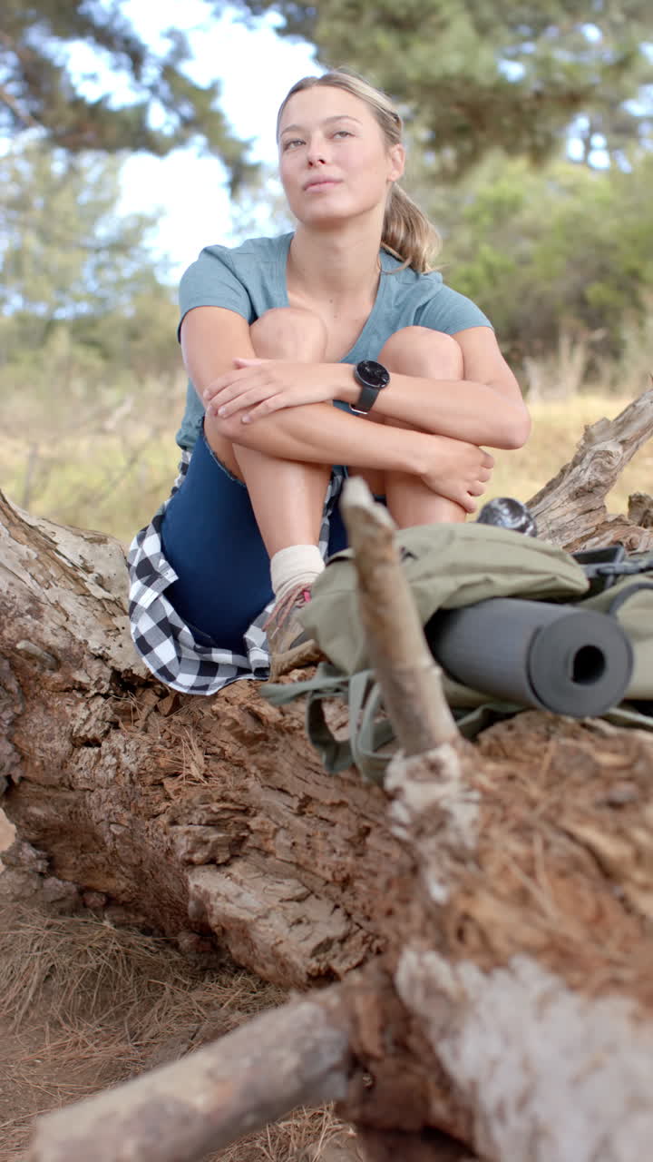 Vertical video: Hiking outdoors, woman sitting on fallen tree with backpack and yoga mat