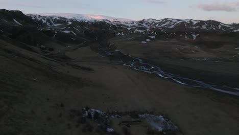 Vista-Aérea-Del-Paisaje-De-Un-Río-Que-Fluye-Por-Los-Picos-De-Las-Montañas-Con-Nieve-Derretida,-En-Islandia,-Al-Atardecer