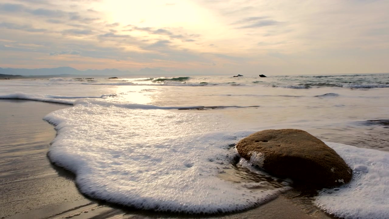 Luz dourada do nascer do sol refletida na areia molhada da praia enquanto pequenas ondas espumosas avançam.