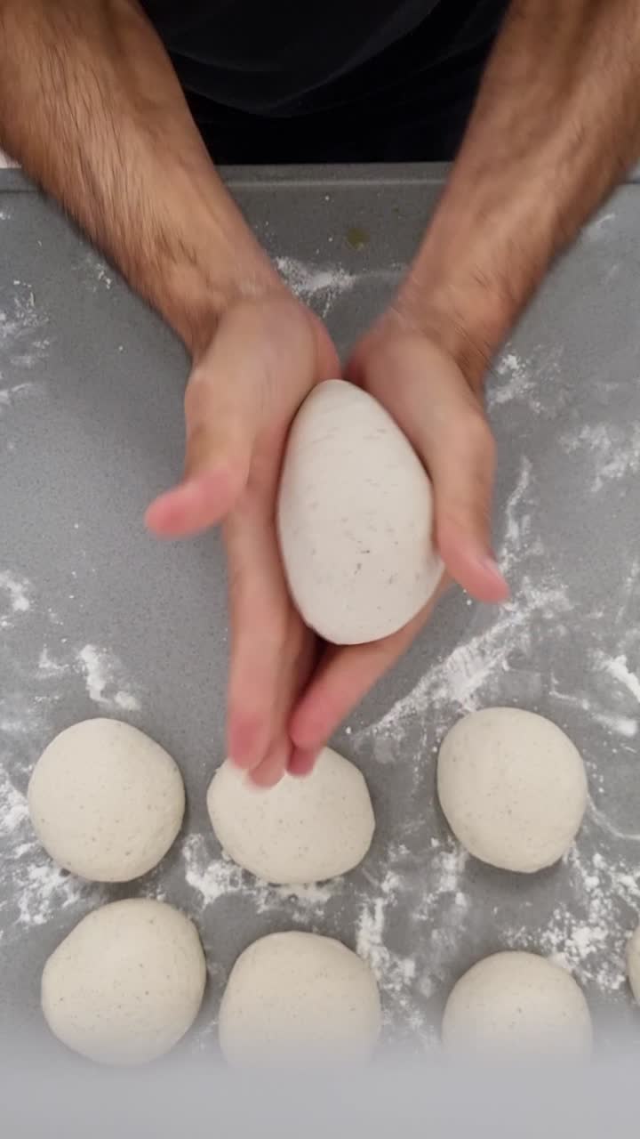 Mãos de pizzaiolo italiano criando bolas de massa de massa fermentada a frio usando o método de dobrar e bolear, filmado em ângulo lateral baixo, vídeo vertical.