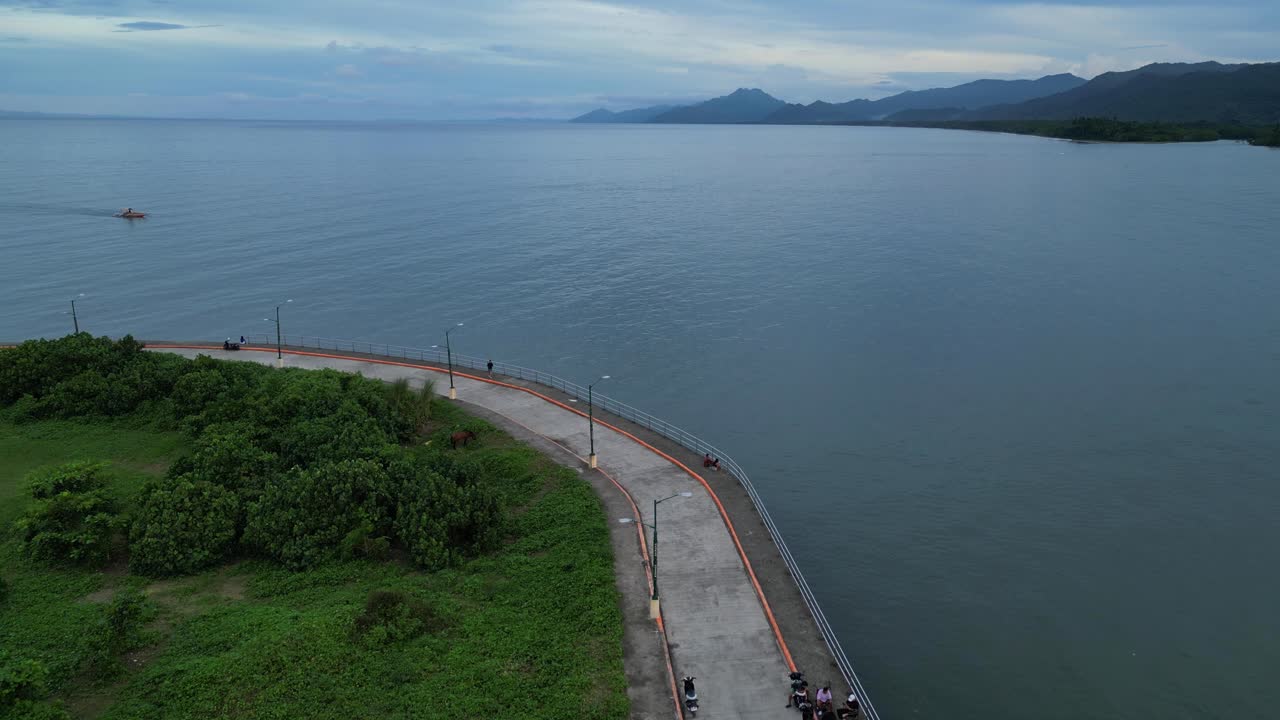 A dolly-in drone shot along a scenic coastal walkway curving beside calm water, with people strolling and biking against a backdrop of mountains and clouds in Mauban Port, Quezon Province Philippines