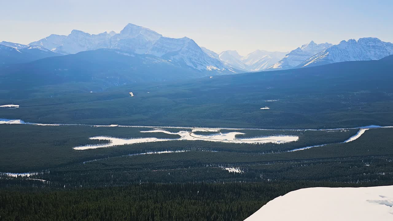 Banff, Canada: Tog bevæger sig gennem et storslået vinterligt bjerglandskab med sneklædte tinder, frosne flodlejer og tæt stedsegrøn skov, der skaber en naturskøn udsigt over vildmarken.