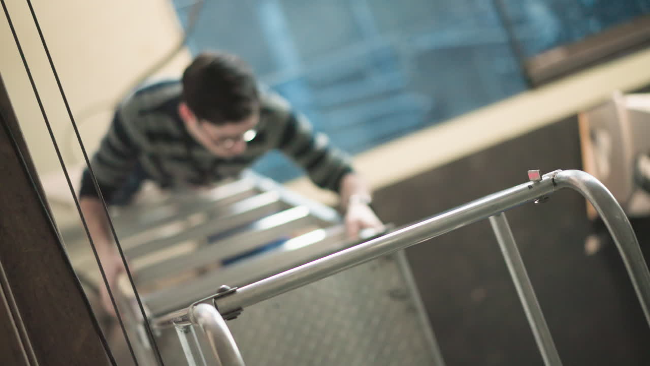 Overhead view of young male employee with glasses wearing striped sweater climbing metal staircase in workspace, gripping handrails, demonstrating upward progress, effort, safety, determination