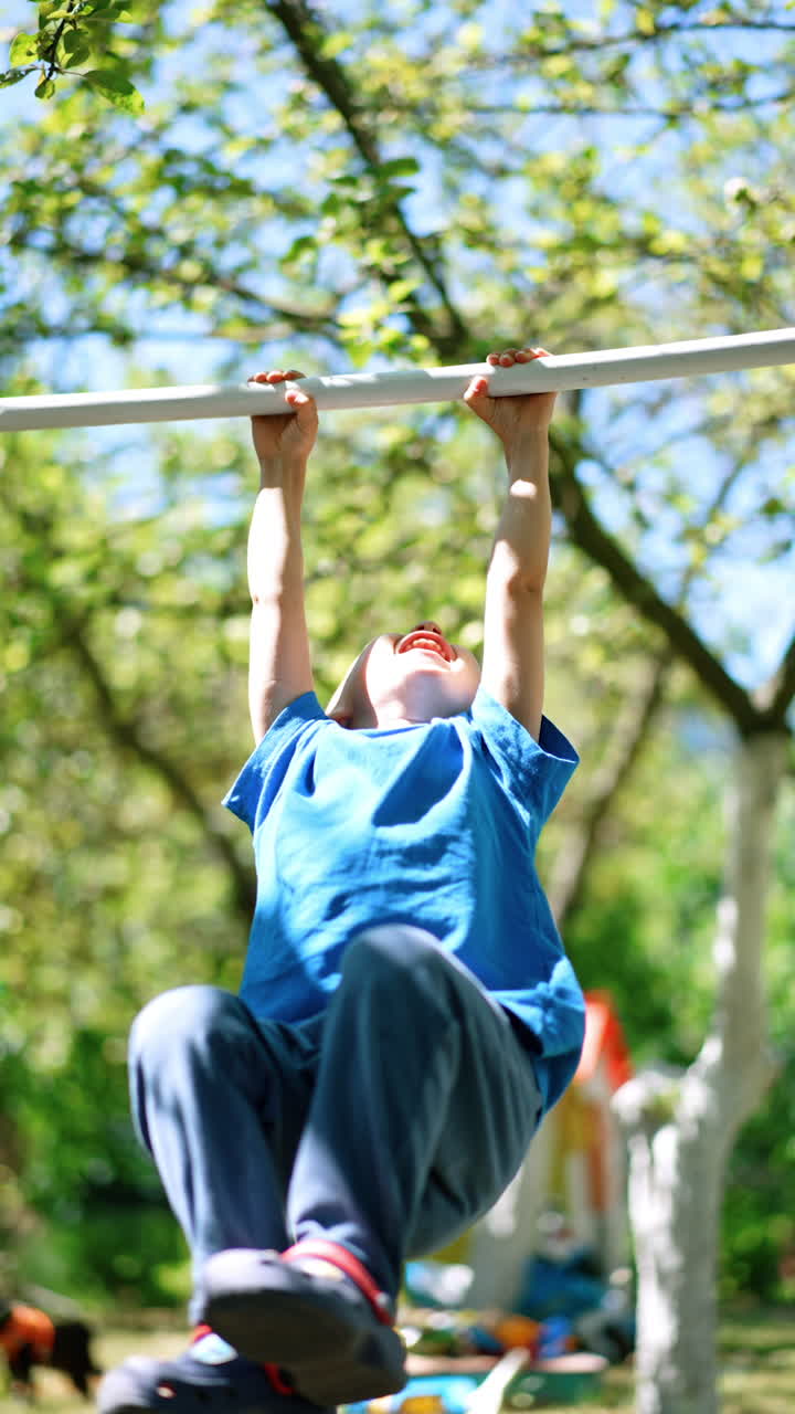 Smiling Caucasian boy stands outdoors holding hands on the horizontal bar. Kid rises feet and waves on the bar. Vertical video.