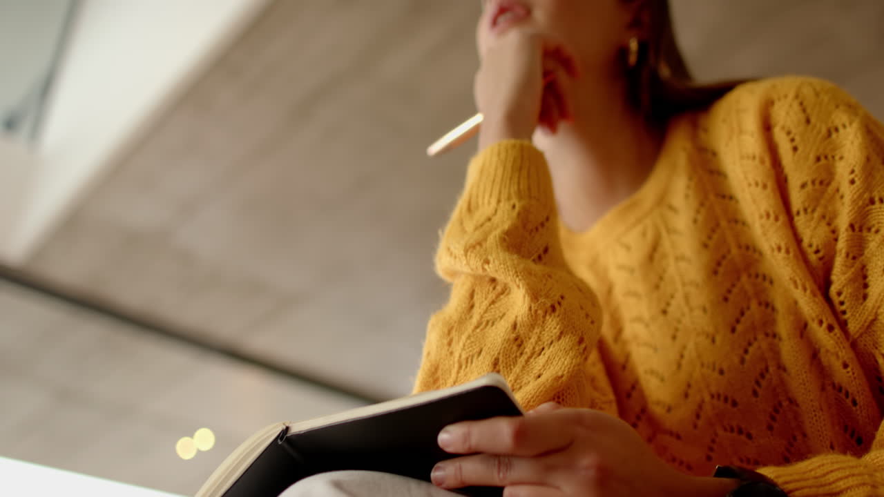 Holding notebook, woman in yellow sweater thinking and writing notes, at home, copy space