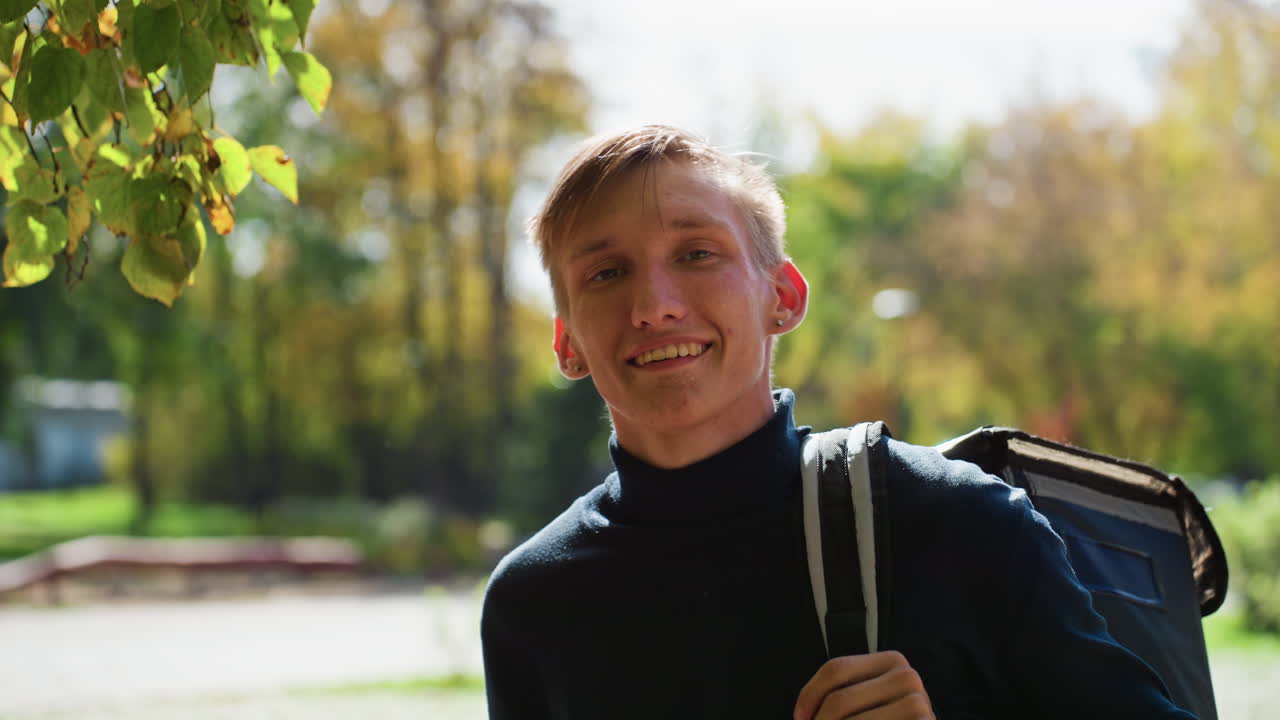 Mladý běloch s batohem venku projevuje úlevu po doručovací trase, sluncem zalitý park v pozadí, podzimní listí, ležérní černý svetr, měnící se výrazy od únavy po úsměv, nesoucí termo tašku