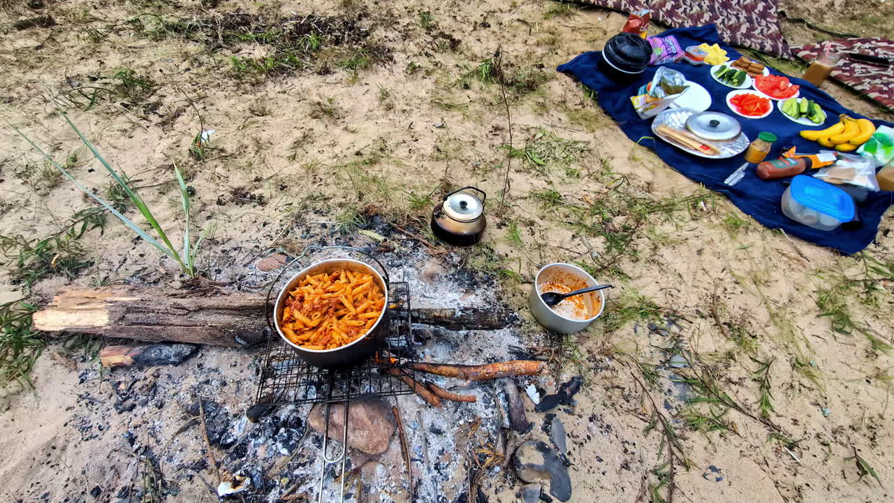 Food cooking over a fire pit with a picnic spread on sand nearby. Tallinn, Estonia