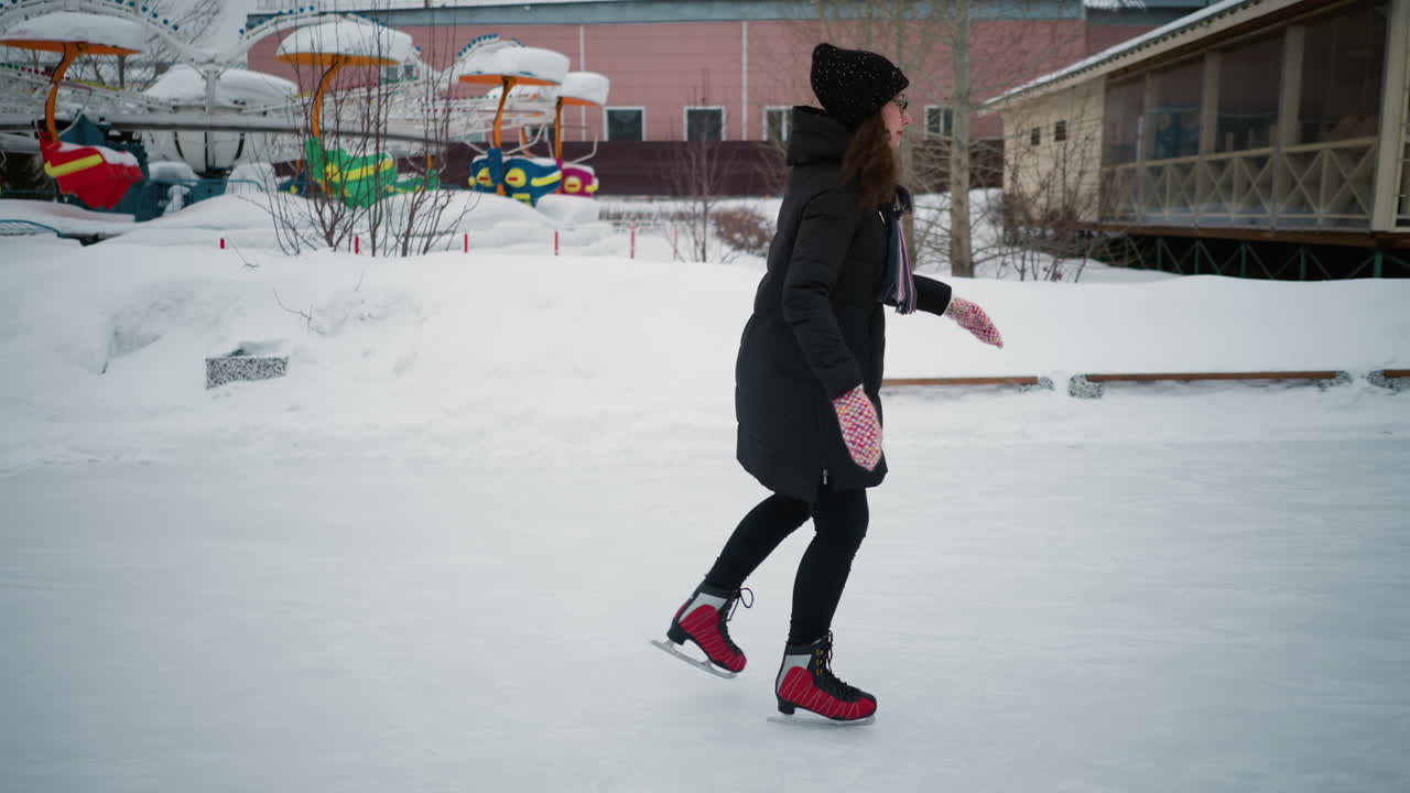 Lady skating on frozen outdoor rink in winter, wearing black coat, knit hat, striped scarf, red skates, and colorful gloves, balancing gracefully on ice with snowy background and amusement rides visible