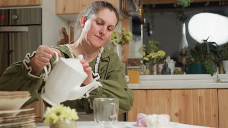 housewife dressed in green top and white apron carefully pouring herbal tea into transparent glass cup at cozy kitchen table with flowers, pastries, and vintage book during peaceful morning routine