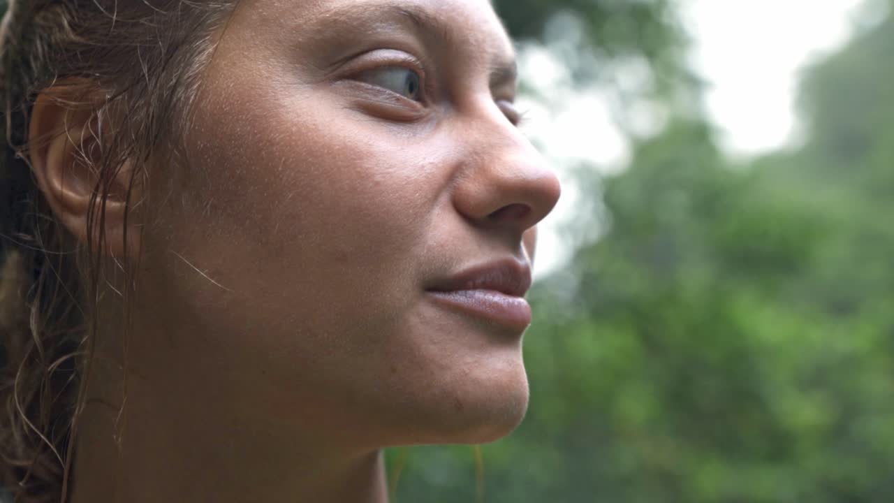 A close-up portrait of a woman with a genuine smile and wet hair, looking up and to the side, with the blurred green foliage of Rio Celeste's rainforest in the background