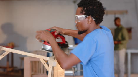 african american man wearing safety goggles sanding wooden frame in woodworking studio, with sander