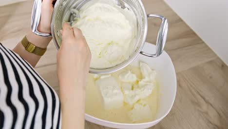 top-down shot shows unrecognizable person spooning mascarpone from pot into bowl with whipped egg yolk and powdered sugar for tiramisu cream, wooden surface beneath, real time, static camera