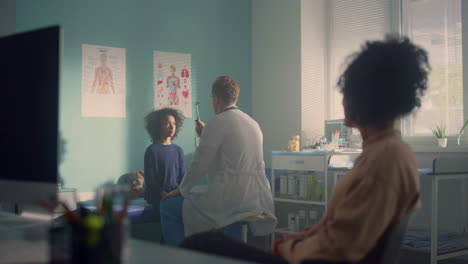 Médico-Revisando-El-Reflejo-Visual-De-La-Niña-En-El-Consultorio.-Mamá-Africana-Esperando-Los-Resultados-De-Las-Pruebas.