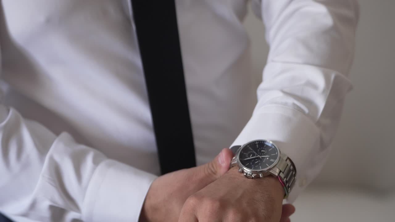 primer plano de un hombre ajustando un reloj de muñeca mientras lleva una camisa blanca y una corbata negra