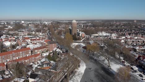 Städtische-Verschneite-Winterszene-Aus-Der-Luft-Mit-Leuten,-Die-Entlang-Des-Gebogenen-Zugefrorenen-Kanals-Eislaufen,-Der-Durch-Die-Niederländische-Stadt-Zutphen-Führt,-Mit-Schatten-Von-Kargen-Bäumen-Und-Ehemaligem-Wasserturm-Im-Hintergrund