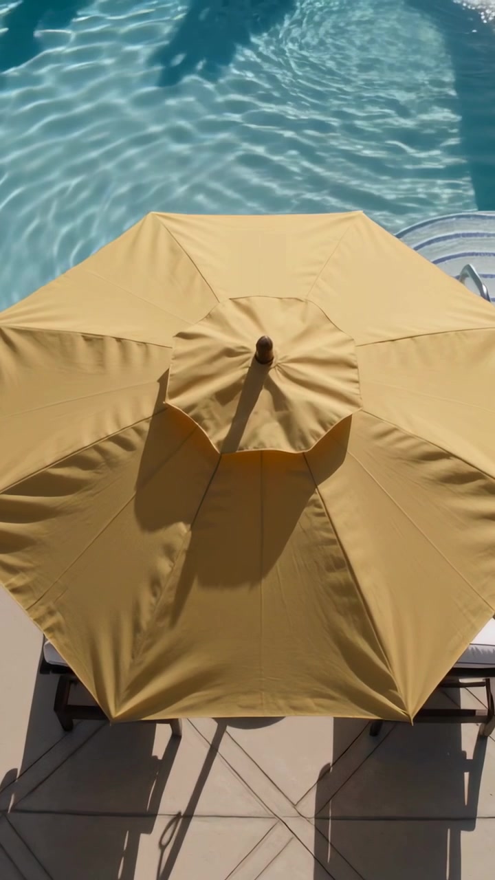 Video verticale: Panoramica della telecamera su un ombrellone giallo che ripara delle sedie a bordo piscina, con la brezza che increspa l'acqua.