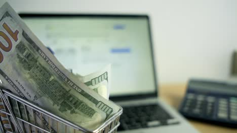billetes de dólar dentro de un carrito de compras juguete al lado de la calculadora portátil en el espacio de trabajo de la oficina en casa, desplazamiento de comercio electrónico, notas verdes americanas físicas