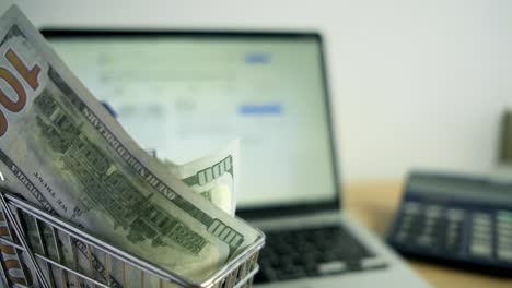 dollar bills inside a shopping cart toy next to calculator laptop in home office workspace, scrolling e-commerce, physical american green notes