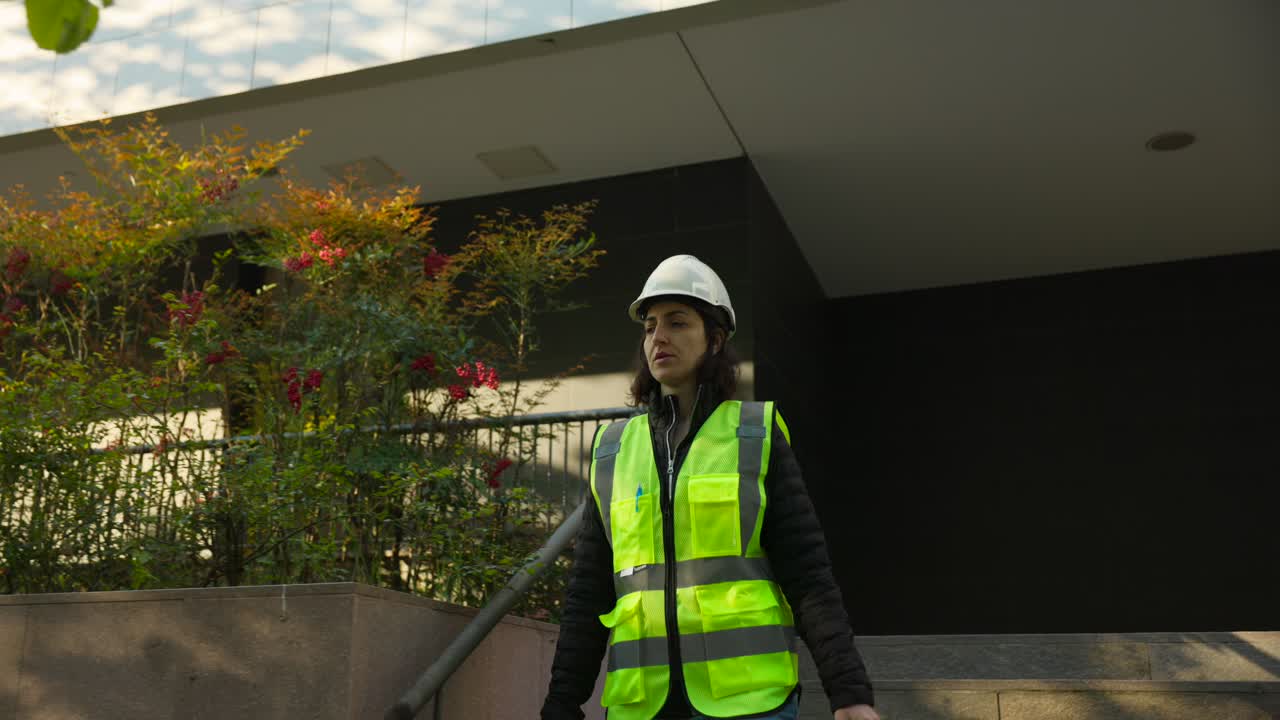 Ingegnera con giubbotto di sicurezza e casco lavora tenendo una custodia per drone mentre scende le scale di un edificio.