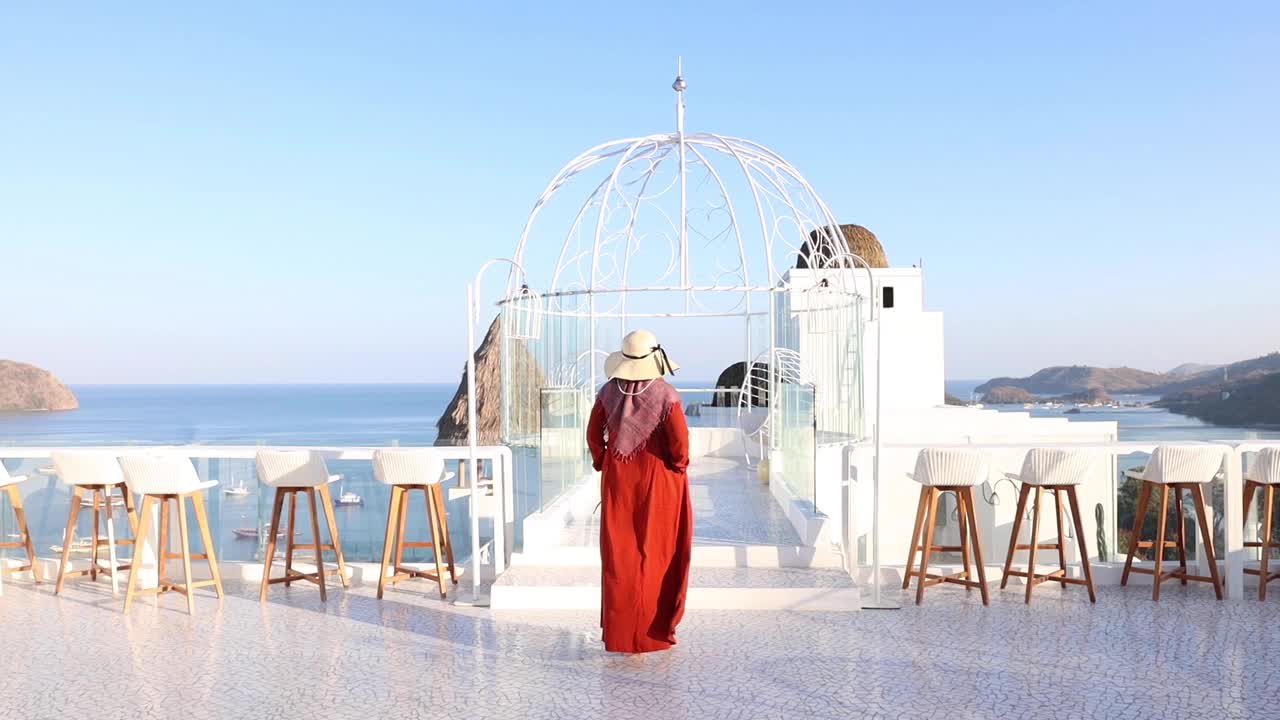 주황색 드레스와 해변 모자를 쓴 이슬람 여성의 뒷모습이 푸른 하늘 아래 걸어가고 있다