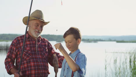 grandfather standing with his teen grandson at the lake shore, talking and holding a fish on the rod