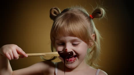 happy little kid girl licking melted chocolate, child eating, enjoying unhealthy sweet food indoors