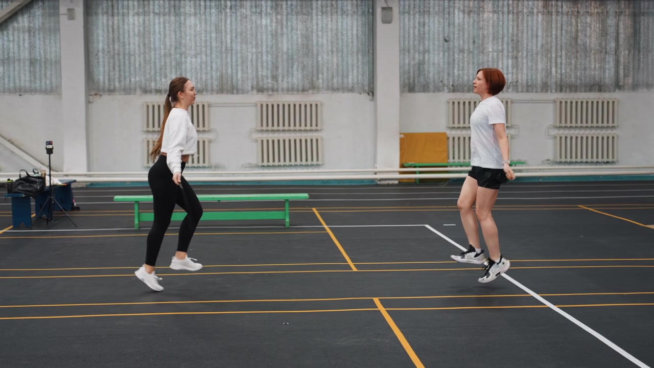 Two women doing badminton warmup routine indoor, synchronized jumps and footwork on lined court, playful coordination and cardio preparation with rackets and shuttlecock, focus on timing and balance