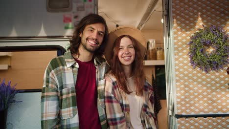 Portrait-of-a-happy-couple,-a-brunette-guy-with-stubble-in-a-Green-checkered-shirt-in-a-red-T-shirt-stands-near-his-brunette-girlfriend-in-a-brown-hat-and-checkered-shirt-Near-his-decorated-trailer-during-a-picnic-in-a-camp-outside-the-city-in-the-summer