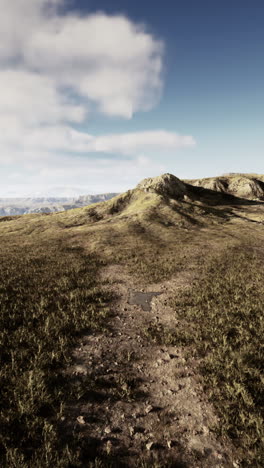 a grassy field with a dirt path leading up a hill, with mountains in the distance and a blue sky with clouds