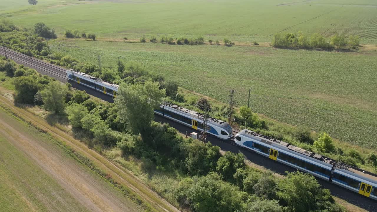 A modern passenger train moves along dual railway tracks through a lush green rural landscape. The scene captures the harmony between transportation and nature, with the train cutting through fields.