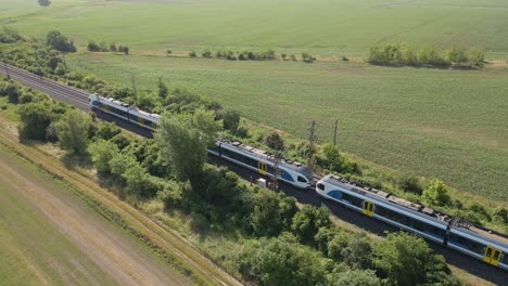 a modern passenger train moves along dual railway tracks through a lush green rural landscape. the scene captures the harmony between transportation and nature, with the train cutting through fields.