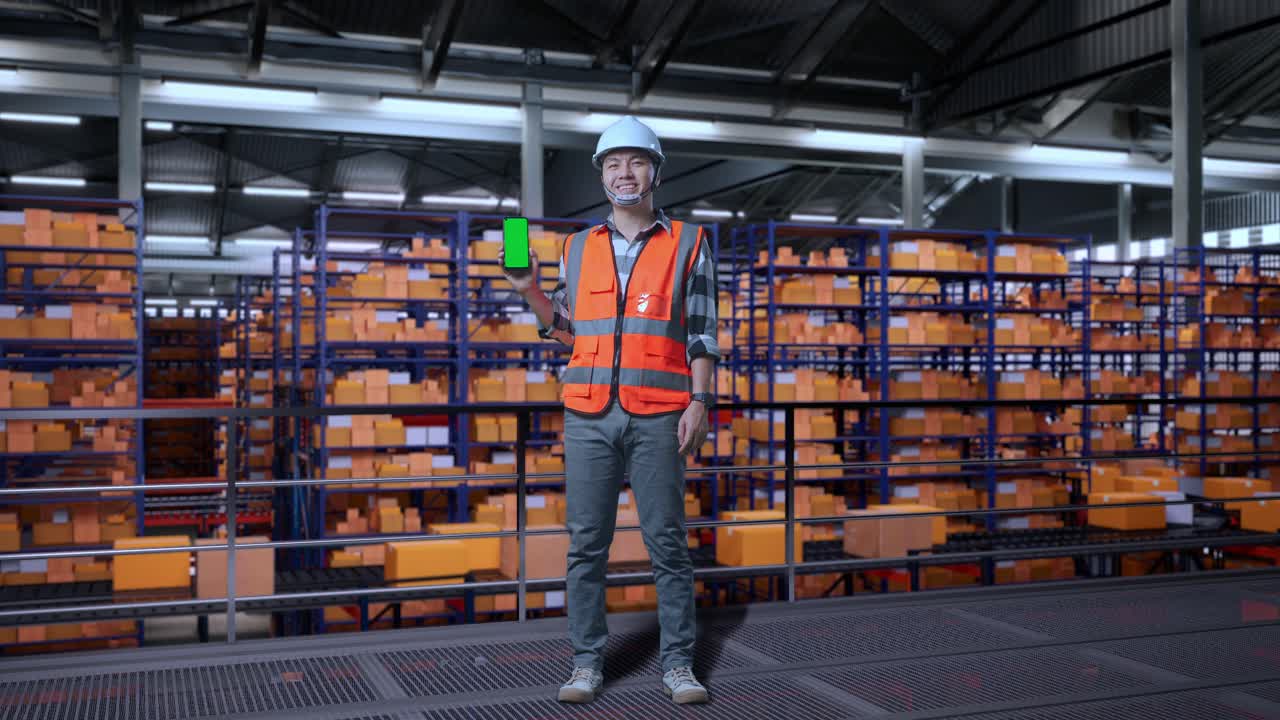 cuerpo lleno de ingeniero masculino asiático con casco de seguridad de pie en el almacén con estantes llenos de bienes de entrega. sonriendo y mostrando pantalla verde teléfono inteligente a la cámara en el almacenamiento