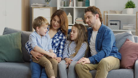 Happy-Family-Sitting-On-The-Cozy-Couch-In-The-Living-Room-And-Talking-With-Their-Small-Cute-Kids-Who-Sitting-On-Their-Knees-And-Between-Them