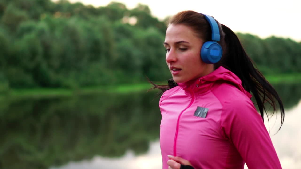 una chica con una chaqueta rosa y pantalones negros corre cerca del río con auriculares preparándose para el maratón