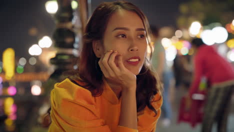 Woman-in-yellow-Ao-Dai-looking-pensive-at-night-in-Hoi-An,-vibrant-street-lights-in-background,-close-up