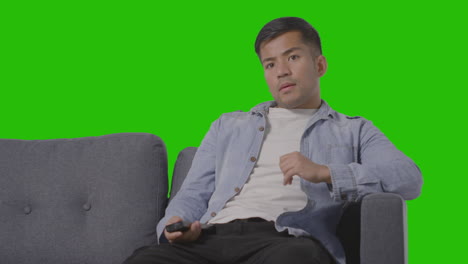 studio shot of young man sitting on sofa with remote control flicking through tv channels against green screen