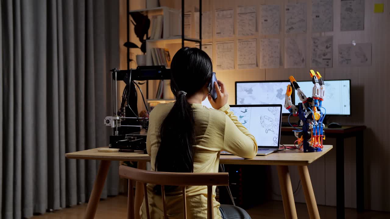 십대 아시아 소녀가 3d 프린팅을 통해 집에 있는 노트북에서 사이보그 손을 디자인하는 동안 스마트폰으로 대화하는 뒷면