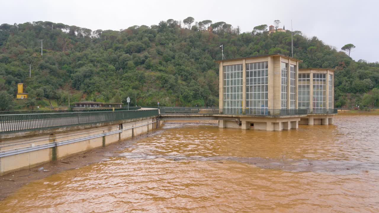 Un fiume in piena di Sau pantano che esonda, gennaio 2026, con acqua marrone fangosa che scorre impetuosamente contro una diga e una torre di controllo. Cambiamento climatico e condizioni meteorologiche estreme che causano inondazioni
