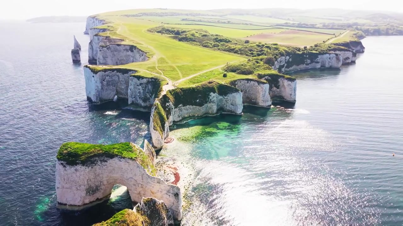 Imágenes aéreas de dron cenitales de Old Harry Rocks en la costa de Dorset en Inglaterra, mostrando acantilados de tiza blanca, cuevas marinas, senderos verdes en la cima del acantilado, agua turquesa poco profunda y mar azul en calma